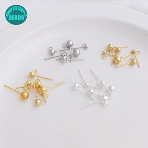 May include: A collection of gold and silver earring posts with small round balls at the top. The earring posts are arranged in three groups on a white surface.