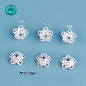 May include: Six silver-colored, flower-shaped bead caps. Each cap has a detailed, openwork design with a central hole. The text "S13 8.0mm" is visible. The caps are arranged on a light blue background. The logo "Anny DIY Supply Beads" is in the top left corner.