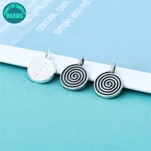 Pode incluir: Três pingentes espirais prateados. Cada pingente é redondo com um design espiral em relevo e uma pequena argola na parte superior para prender a uma corrente ou outras joias. Os pingentes são exibidos em uma superfície azul clara.