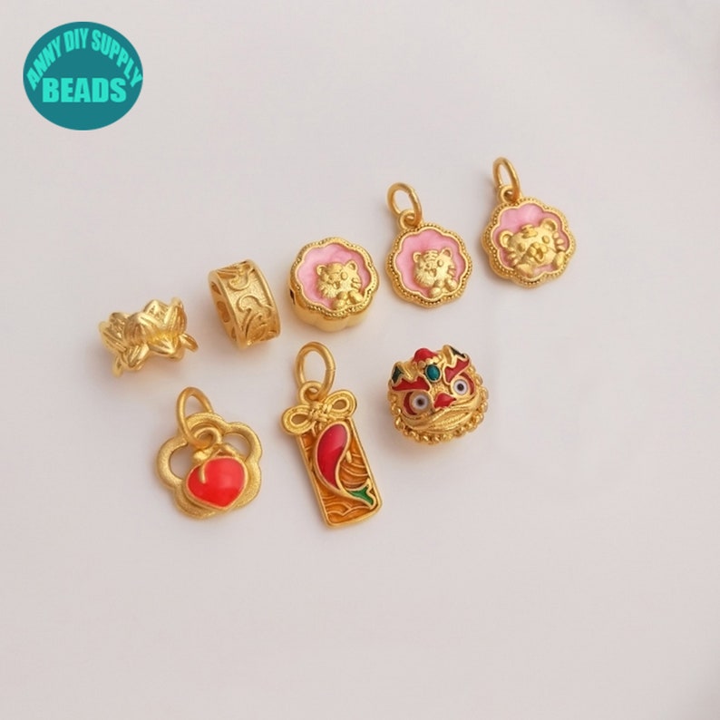 May include: A collection of gold-colored charms and beads. The charms include a lotus flower, a patterned cylinder, pink-rimmed tiger pendants, a red heart-shaped charm, a red and gold pendant, and a detailed lion head bead. The text "ANNY DIY SUPPLY BEADS" is in a circle.