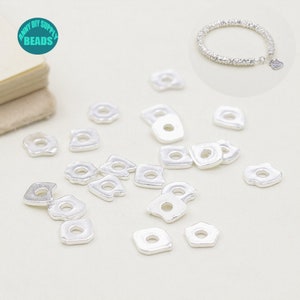 Könnte beinhalten: Silberne Sprungringe, eine Art von Schmuckzubehör, sind auf einer weißen Oberfläche verstreut. Die Sprungringe sind klein und haben eine quadratische Form mit einem runden Loch in der Mitte. Im Hintergrund befindet sich ein silbernes Armband.