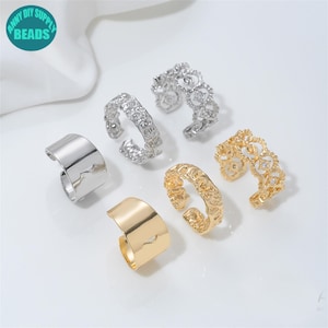 Könnte beinhalten: Eine Sammlung von sechs offenen Ringen in Silber- und Goldtönen. Das Set enthält zwei glatte, polierte Bänder und vier Ringe mit komplexen, strukturierten oder filigranen Mustern. Jeder Ring verfügt über eine verstellbare Öffnung.