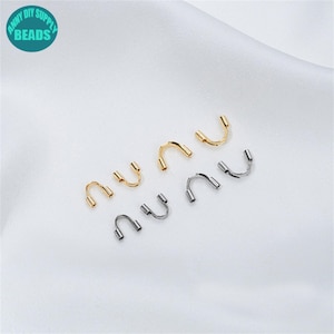 Op de afbeelding: Een verzameling goud- en zilverkleurige U-vormige sieradenonderdelen. De onderdelen hebben een gebogen ontwerp met twee kleine, cilindrische pinnen aan elk uiteinde. De achtergrond is een witte, zijdeachtige stof. De tekst "ANNY DIY SUPPLY BEADS" is inbegrepen.