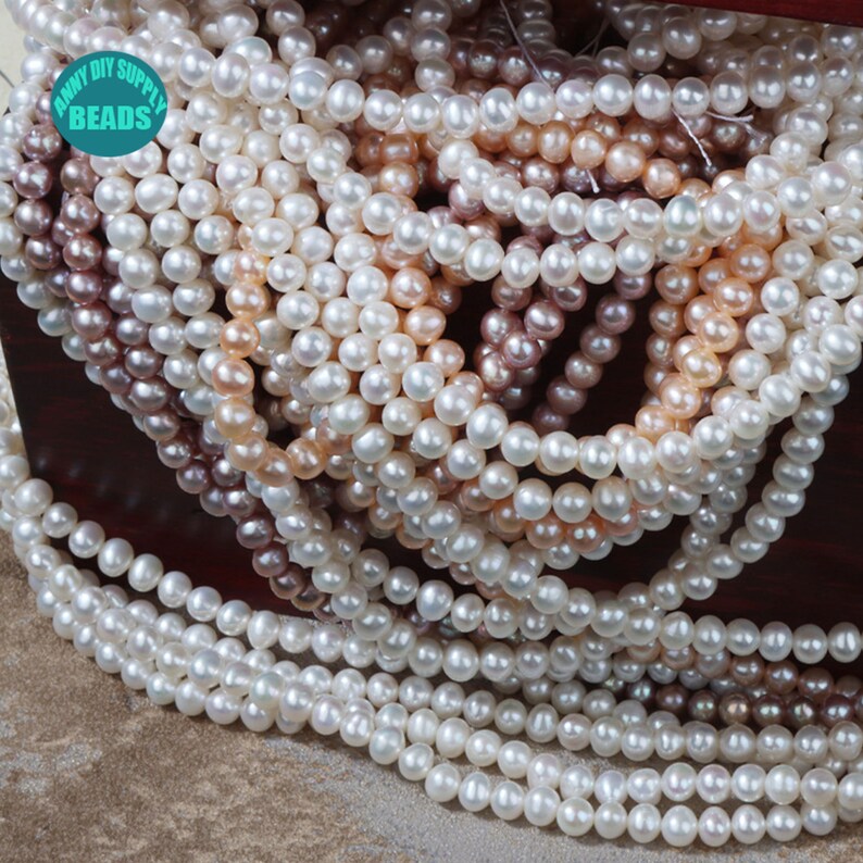 May include: A close-up of a pile of white, cream, and pink pearl beads. The beads are round and smooth, and they are arranged in a random pattern. The beads are in a wooden box with the text "ANNY DIY SUPPLY BEADS" on the front.