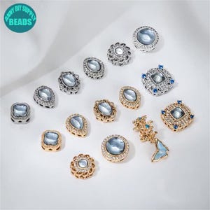 Pode incluir: Uma variedade de contas decorativas em várias formas, incluindo redondas, ovais e quadradas. As contas apresentam uma pedra central azul clara rodeada por engastes de metal prateado ou dourado, algumas com detalhes de cristal transparente. O texto "ANNY DIY SUPPLY BEADS" está incluído.