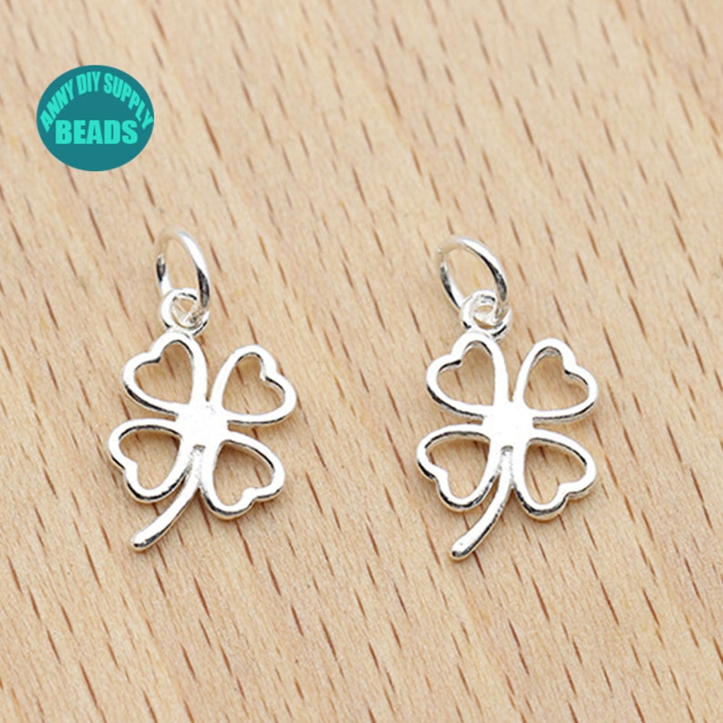 May include: Two silver four-leaf clover charms with a jump ring for attaching to jewelry. The charms are on a light brown wooden surface.
