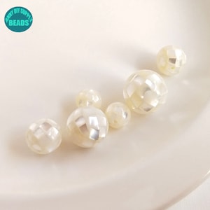 Könnte beinhalten: Eine Sammlung von sechs weißen, irisierenden runden Perlen mit einem facettierten Design. Die Perlen sind in einer Reihe auf einer weißen Oberfläche angeordnet.