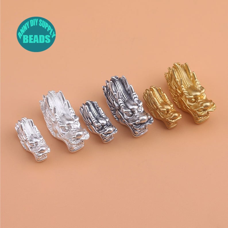 May include: Five silver and gold dragon head charms. The charms are in different sizes and are arranged in a row from smallest to largest. The charms are all facing the right side of the image.