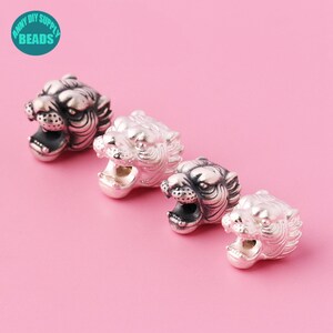 Puede incluir: Cuatro cuentas plateadas con forma de cabeza de tigre con la boca abierta, dispuestas en diagonal sobre un fondo rosa. Las cuentas tienen rasgos faciales detallados y probablemente son para hacer joyas. El texto "ANNY DIY SUPPLY BEADS" está en un círculo turquesa.