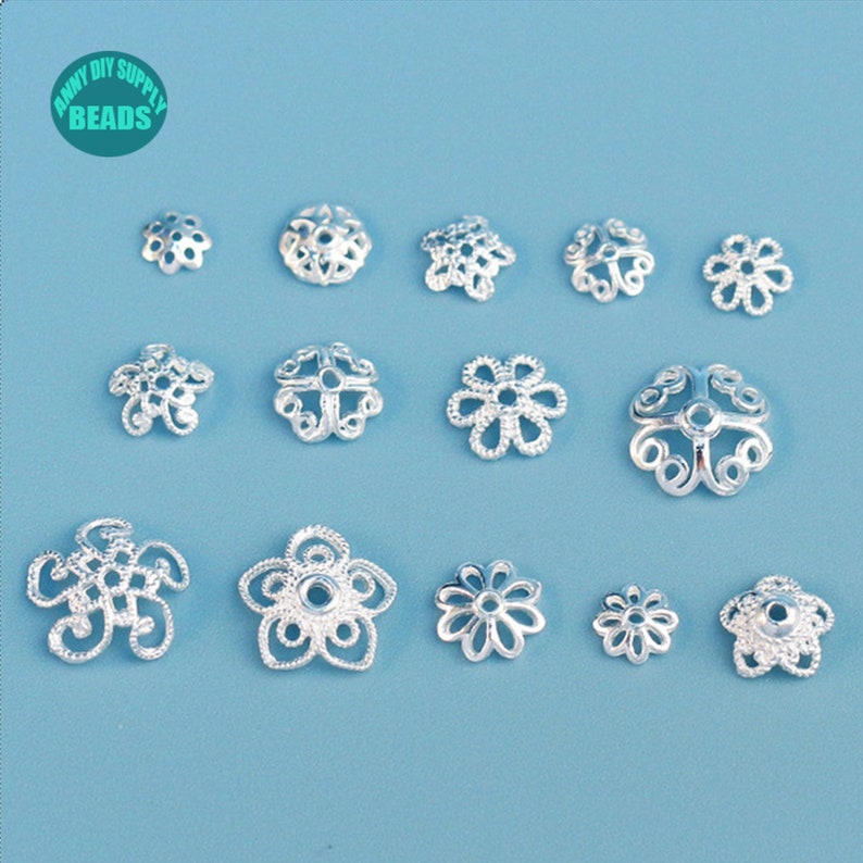 May include: An assortment of silver-colored, filigree-style bead caps in various floral designs. The bead caps are arranged on a light blue background. The image includes the text "ANNY DIY SUPPLY BEADS" in a circular logo.