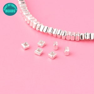 Könnte beinhalten: Silberfarbene quadratische Perlen mit einem Loch in der Mitte sind auf einer rosa Oberfläche verstreut. Ein Strang ähnlicher Perlen befindet sich im Hintergrund. Das Bild enthält ein Logo mit der Aufschrift "ANNY DIY SUPPLY BEADS".