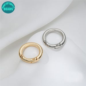 Könnte beinhalten: Zwei Metall-Federringverschlüsse. Einer ist goldfarben, der andere silberfarben. Beide sind kreisförmig mit einem Scharnieröffnungs- und Schließmechanismus. Die Verschlüsse sind für die Schmuckherstellung oder andere Bastelprojekte bestimmt. Der Text "ANNY DIY SUPPLY BEADS" ist enthalten.