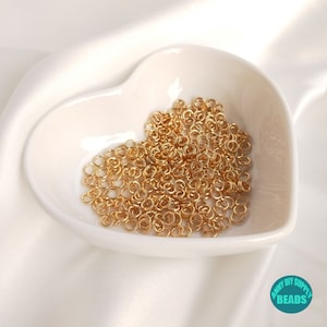 May include: A heart-shaped white ceramic dish filled with a large quantity of small, gold-colored jump rings. The rings are perfect for jewelry making and crafting.
