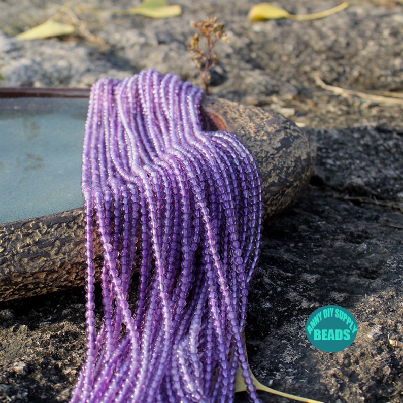K&ouml;nnte beinhalten: Nahaufnahme einer Schnur aus violetten Glasperlen. Die Perlen sind klein und rund und haben einen leichten irisierenden Schimmer. Die Perlen sind in einem lockeren Haufen angeordnet und liegen auf einem St&uuml;ck rauer, brauner Rinde.