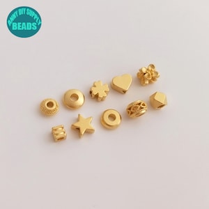 Op de afbeelding: Assortiment van goudkleurige metalen kralen in verschillende vormen, waaronder een ster, een hart, een bloem, een klavertjevier en cirkels. De kralen zijn klein en perfect voor het maken van sieraden.