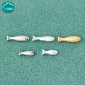 Puede incluir: Cinco dijes de metal con forma de pez, en plata y oro. Los dijes están dispuestos sobre una superficie turquesa. Los peces tienen aletas y detalles de ojos. La esquina superior izquierda de la imagen tiene el texto "ANNY DIY SUPPLY BEADS".