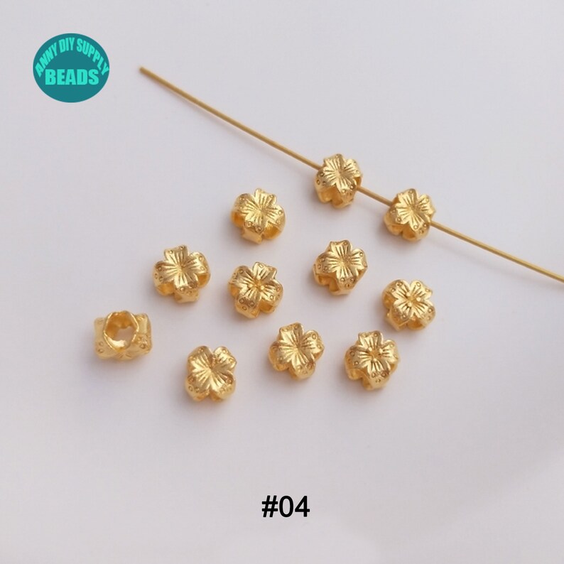 May include: Gold-toned metal flower-shaped bead caps. There are 11 bead caps in the image. The bead caps are arranged in a scattered pattern on a white surface. The image also includes a gold-toned wire.