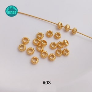May include: A close-up of a group of gold-colored metal beads with a textured surface. The beads are arranged in a scattered pattern on a white surface. The beads are approximately 5mm in diameter. #03