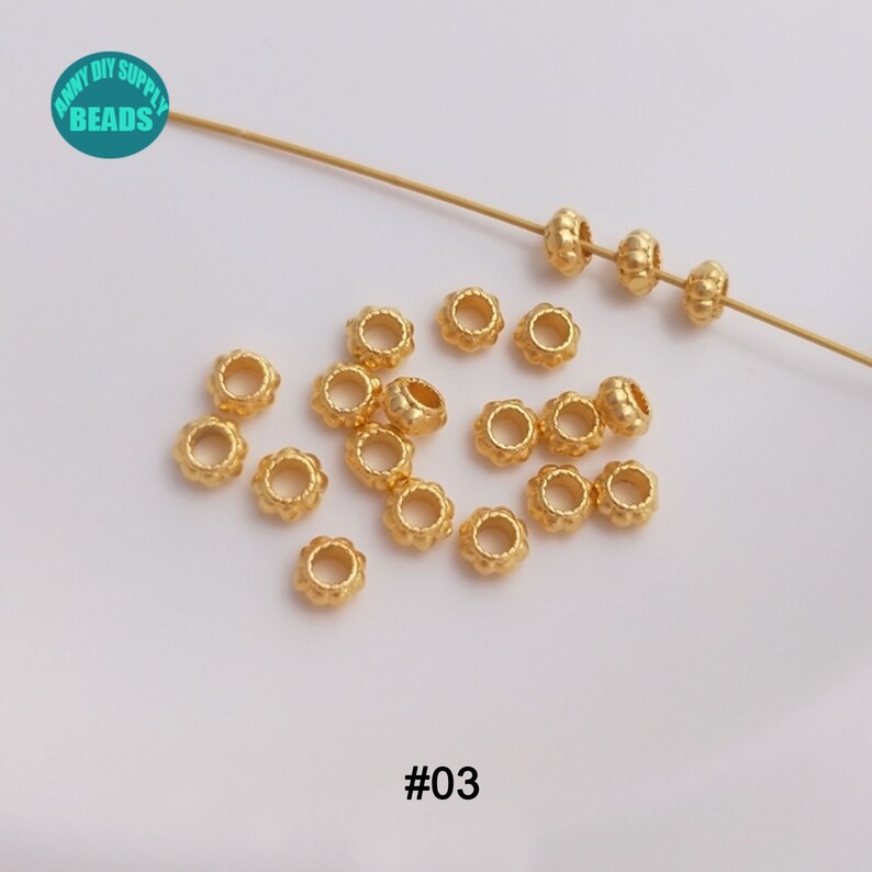 May include: A close-up of a group of gold-colored metal beads with a textured surface. The beads are arranged in a scattered pattern on a white surface. The beads are approximately 5mm in diameter. #03