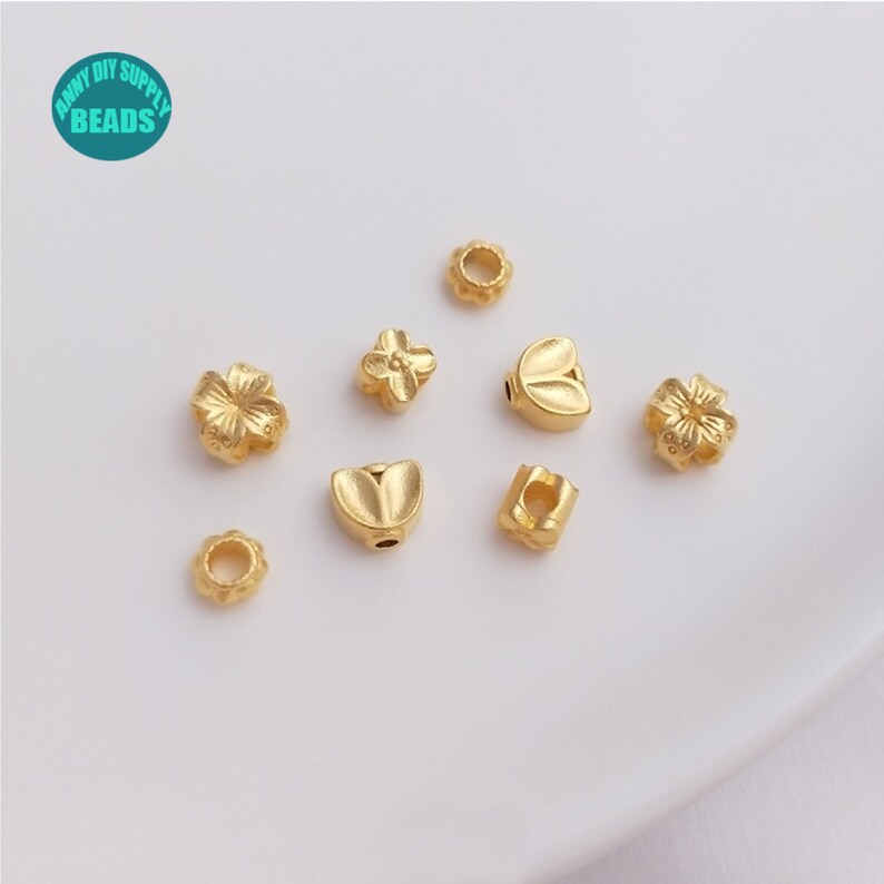 May include: Assortment of gold-toned metal beads with various designs, including flowers, leaves, and a heart. The beads are small and have a shiny finish.