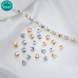 Puede incluir: Anny DIY Supply Beads. Surtido de cuentas cúbicas metálicas en oro y plata, dispersas sobre una superficie blanca. Un hilo de perlas y cuentas cúbicas es visible en el fondo. Las cuentas son para hacer joyas.