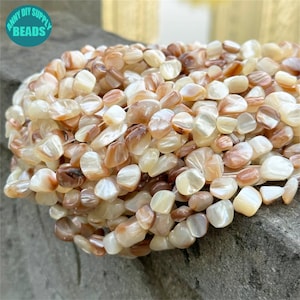 Puede incluir: Una hebra de cuentas de concha marrón y blanca, con una forma ligeramente irregular, plana y de guijarro. Las cuentas están enhebradas en un hilo transparente.