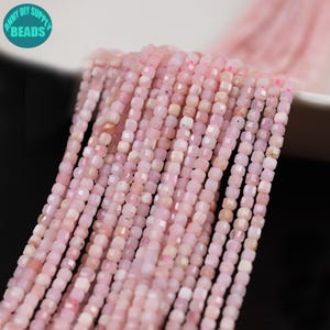 May include: Multiple strands of small, faceted, light pink cube-shaped beads arranged in parallel lines. The beads have a subtle, semi-translucent appearance with slight variations in tone. A circular teal logo with the text ANNY DIY SUPPLY BEADS.