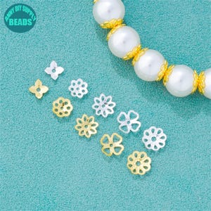 Könnte beinhalten: Eine Auswahl an blütenförmigen Schmuckteilen in Gold- und Silbertönen, auf einer türkisfarbenen Oberfläche angeordnet. Im Hintergrund ist eine Kette aus großen weißen Perlen mit goldenen Akzenten zu sehen. Der Text "ANNY DIY SUPPLY BEADS" ist enthalten.