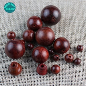May include: A collection of round, dark red wooden beads of various sizes. The beads have a smooth, polished finish and a hole drilled through the centre. The beads are scattered on a textured, beige fabric surface.