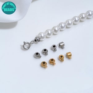 May include: A string of white spherical beads with a silver-toned circular clasp attached to one end, accompanied by four silver-toned and four gold-toned cylindrical crimp beads with side screws, arranged in two rows below the bead strand.