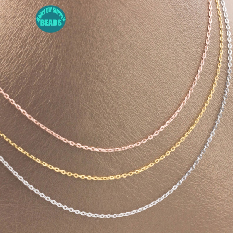 May include: Three chains of different colors: silver, gold, and rose gold. The chains are arranged in a layered pattern on a brown surface.  The chains are all the same style, with a delicate, oval-shaped link.