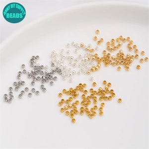 May include: A close-up of a variety of small, round beads in gold, silver, and a light silver colour. The beads are scattered on a white surface.