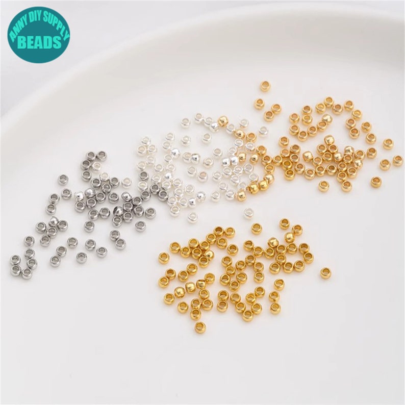 May include: A close-up of a variety of small, round beads in gold, silver, and a light silver colour. The beads are scattered on a white surface.