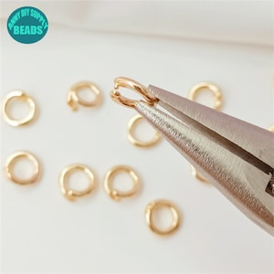May include: Gold jump rings being closed with pliers. The jump rings are small and round, and there are several of them scattered on a white surface.