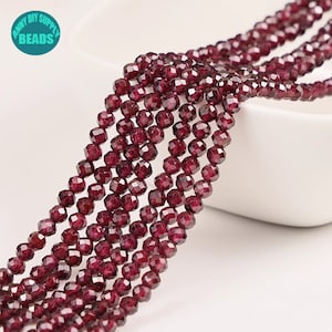 Puede incluir: Una hebra de cuentas de granate rojo oscuro, facetadas y redondas, enhebradas en una superficie blanca. Las cuentas son pequeñas y de tamaño uniforme, creando un aspecto liso y elegante.