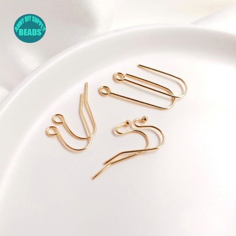 May include: Gold-toned earring hooks, some with a small ball at the end, on a white surface.  The hooks are for jewelry making.
