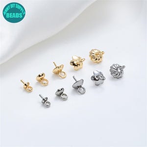 Pode incluir: Uma coleção de achados de joias douradas e prateadas, incluindo pinos de brinco e tampas de contas decorativas. Os achados são dispostos em uma superfície branca, mostrando suas várias formas e designs. A imagem inclui o texto "ANNY DIY SUPPLY BEADS".