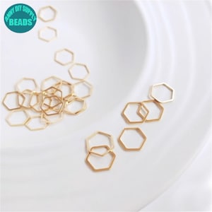 May include: A group of gold-coloured metal hexagonal jump rings. The rings are scattered on a white surface.