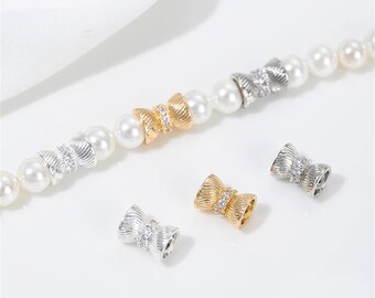 May include: A close-up of a silver and gold bow-shaped bead with clear rhinestones. The bead is part of a pearl necklace.
