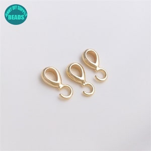 Pode incluir: Três achados de joalheria de metal dourado com um laço e um gancho. Os achados estão dispostos em uma fila sobre uma superfície branca.