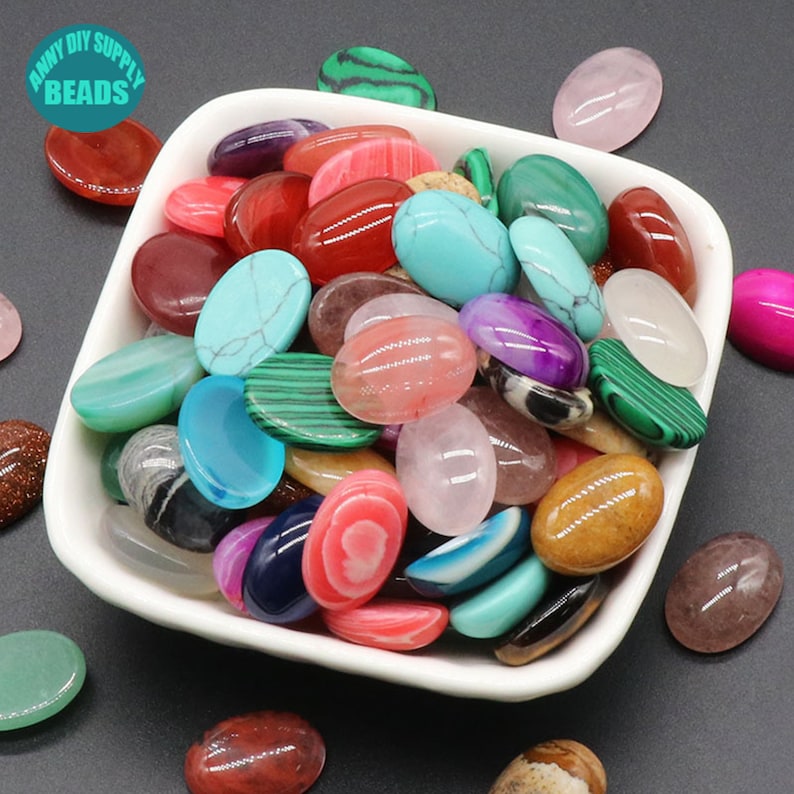 May include: A white bowl filled with a variety of oval-shaped gemstone cabochons in various colors, including red, green, blue, pink, and brown. The cabochons are smooth and polished, and they are perfect for jewelry making or other craft projects.