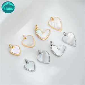 Op de afbeelding: Een set van zeven gouden en zilveren hartvormige bedels met een witte parelmoer inleg. De bedels zijn perfect voor het maken van sieraden en knutselen.