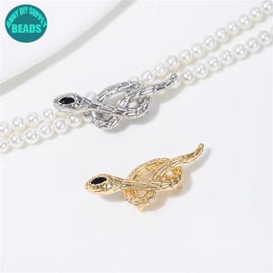 May include: Two snake-shaped brooches, one silver and one gold, with black eyes. The brooches are on a white pearl necklace.