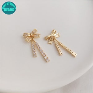 Può includere: Due orecchini dorati a forma di fiocco con accenti lineari pendenti. Un orecchino presenta pietre preziose rotonde e trasparenti lungo gli accenti pendenti. L'altro orecchino ha un design simile, ma con un motivo diverso. Gli orecchini sono su una superficie bianca.