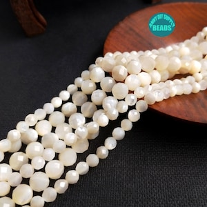 以下が含まれることがあります： 虹色の白い宝石ビーズのストランド。ビーズは丸く、ファセット加工が施されています。ビーズは透明な糸に通されており、木製の表面に表示されています。