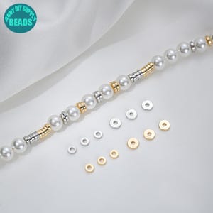 May include: A strand of white pearl beads with gold and silver spacers. The beads are arranged in a line with the spacers in between. There are also a number of small gold and silver jump rings scattered around the beads.