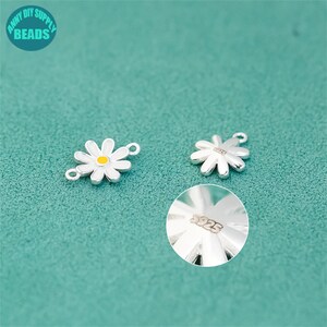 K&ouml;nnte beinhalten: Zwei silberne Anh&auml;nger in Form einer G&auml;nsebl&uuml;mchen mit wei&szlig;en Bl&uuml;tenbl&auml;ttern und gelben Zentren. Ein Anh&auml;nger hat eine Schlaufe zum Befestigen an Schmuck. Der andere Anh&auml;nger hat die Nummer "925" eingestanzt. Der Hintergrund ist t&uuml;rkis und der Text "ANNY DIY SUPPLY BEADS" befindet sich oben links.