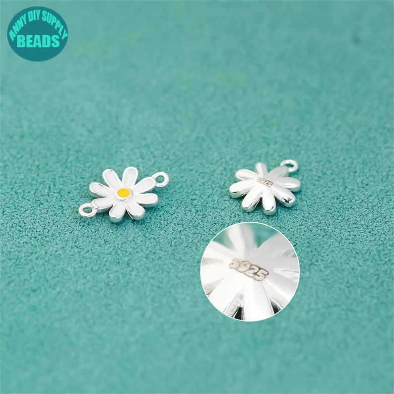 K&ouml;nnte beinhalten: Zwei silberne Anh&auml;nger in Form einer G&auml;nsebl&uuml;mchen mit wei&szlig;en Bl&uuml;tenbl&auml;ttern und gelben Zentren. Ein Anh&auml;nger hat eine Schlaufe zum Befestigen an Schmuck. Der andere Anh&auml;nger hat die Nummer "925" eingestanzt. Der Hintergrund ist t&uuml;rkis und der Text "ANNY DIY SUPPLY BEADS" befindet sich oben links.