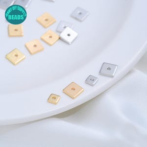 Könnte beinhalten: Eine Sammlung kleiner, flacher, quadratischer Metallperlen in Gold- und Silbertönen, die jeweils ein mittiges Loch aufweisen. Die Perlen sind auf einer glatten, geschwungenen Oberfläche verstreut, mit einem türkisfarbenen runden Logo ANNY DIY SUPPLY BEADS.