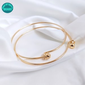 May include: Two gold bangle bracelets with a ball clasp. The bracelets are stacked on top of each other.
